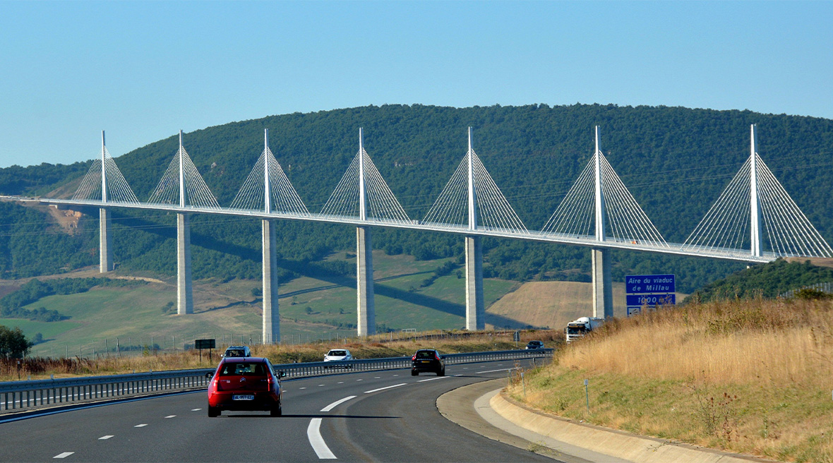 A huge cable span bridge runs over and across a dual carriageway with cars travelling towards it